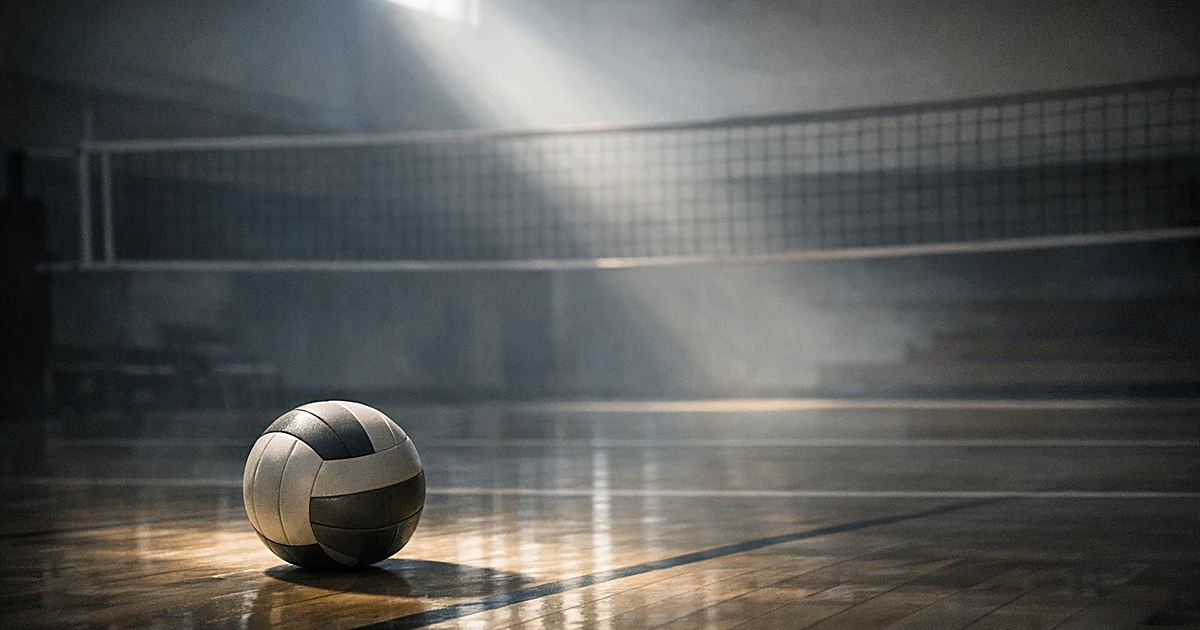 A volleyball sits alone on an empty indoor court with dramatic lighting creating shadows across the wooden floor