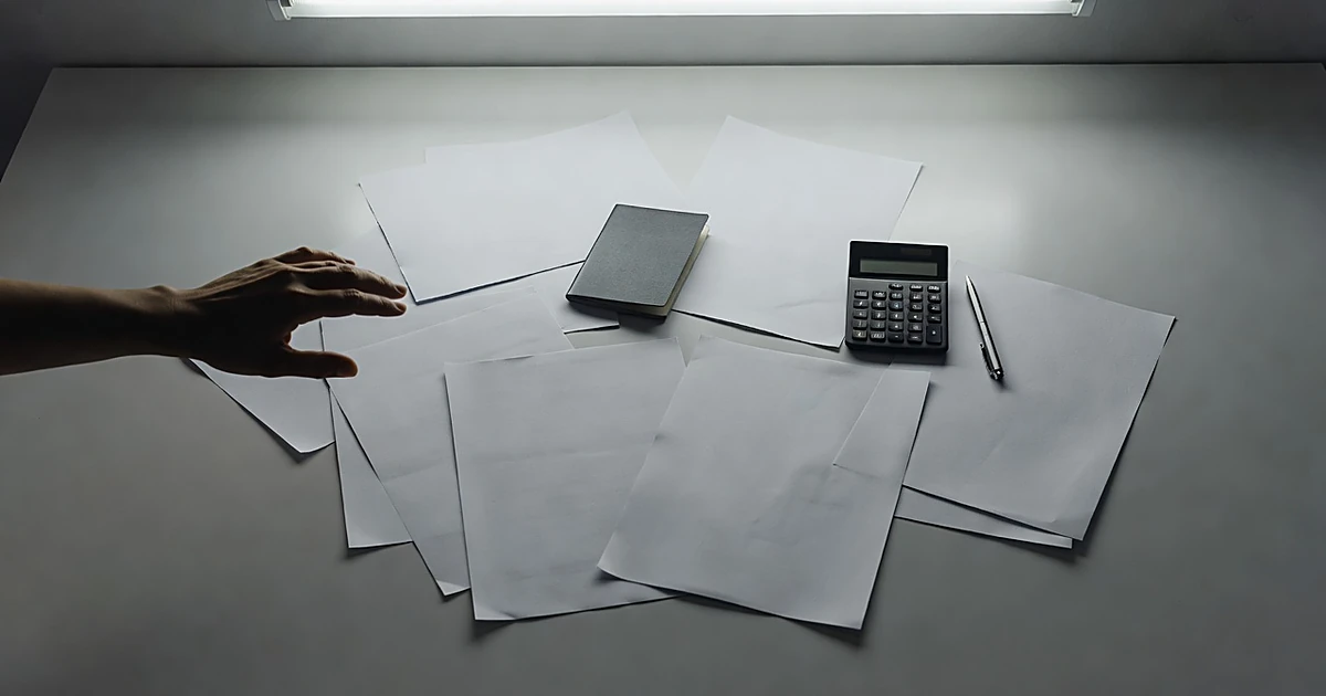 Overhead view of blank tax forms, passport, and calculator on desk with silhouetted hand reaching toward documents
