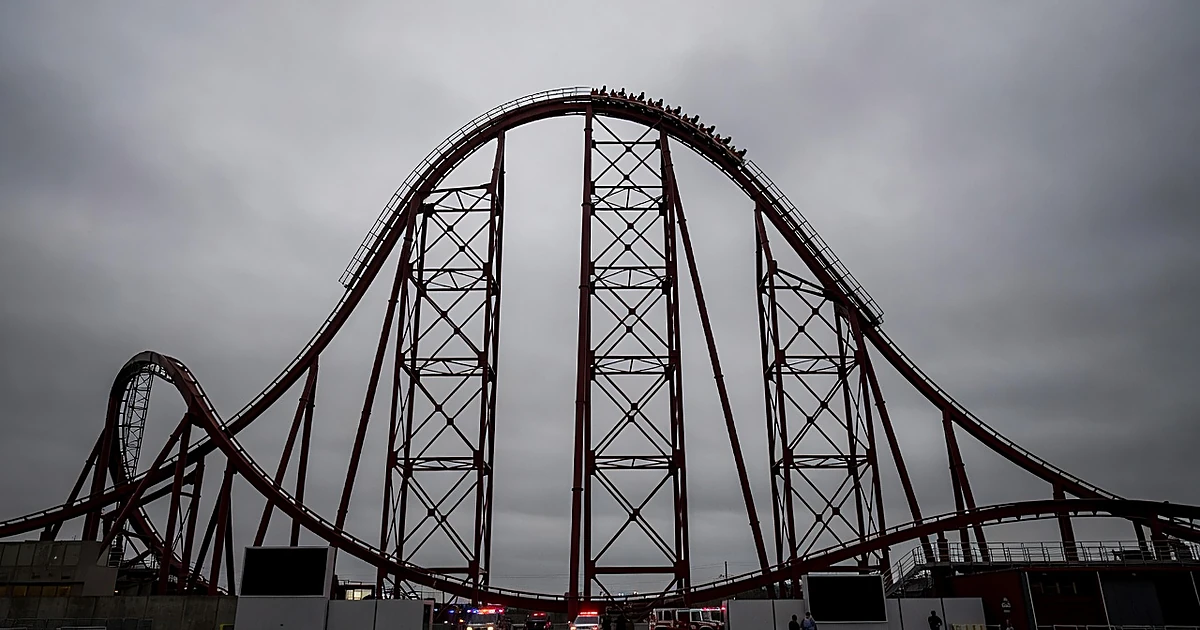 Tall roller coaster structure with emergency vehicles at base, cars stopped near top of track against gray sky