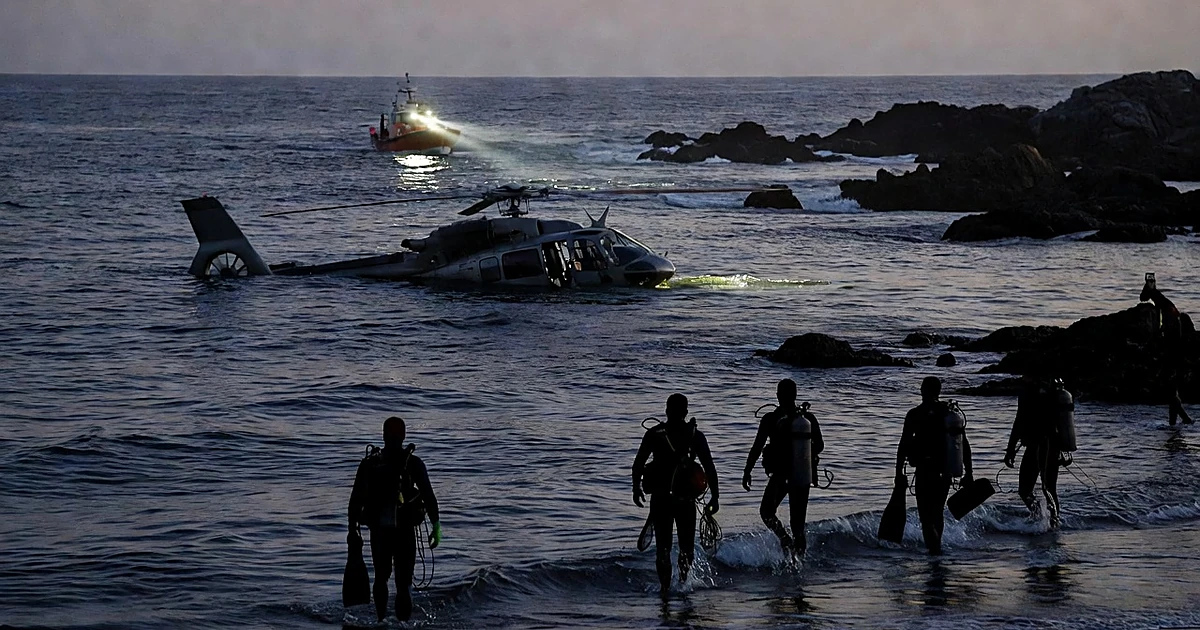 Medical helicopter partially submerged in ocean waters with rescue divers silhouetted in foreground at dusk
